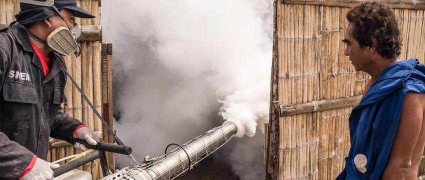 Photo of man in safety gear spraying smoke into a enclosed fence area while another man looks on.