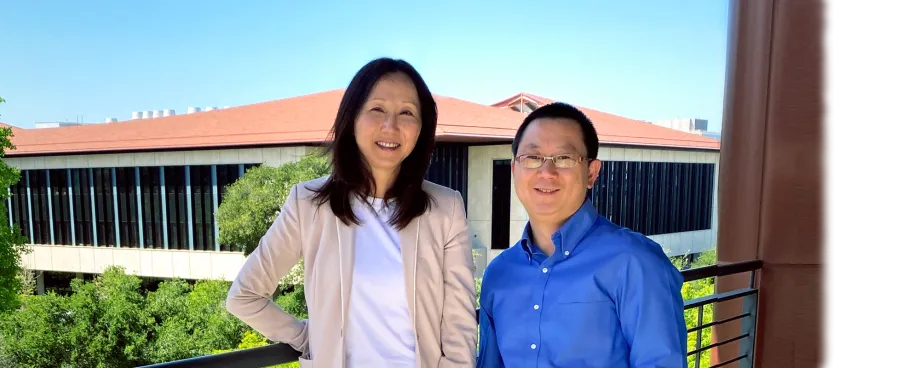 Outdoor photo on a balcony of a smiling Asian female faculty member wearing a pink blazer and a white shirt, with long black hair, and a slightly shorter, smiling male Asian faculty member wearing glasses and a royal blue button up shirt. In the distance behind them are bright green trees and a building with many windows and a red brick roof.
