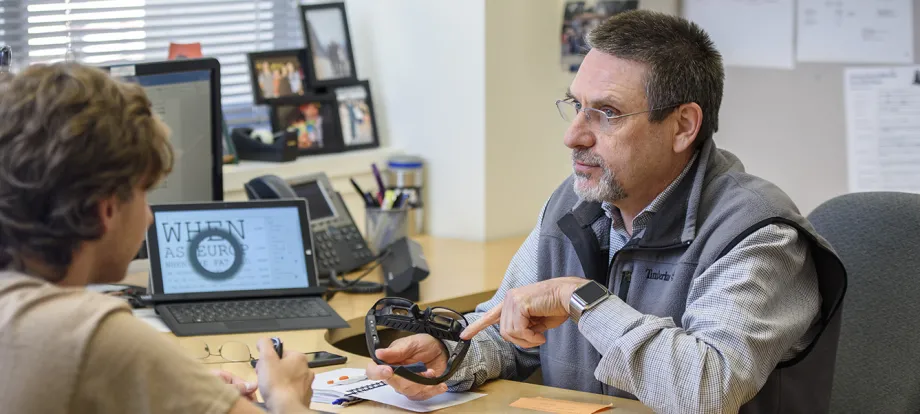 Photo of male PI sitting at table with student, with computer screen displaying readouts, holding a device shaped like safety glasses.