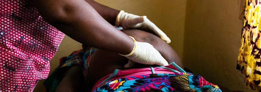 Photo of an African doctor wearing pink scrubs and pale yellow gloves examinign a pregnant African woman.