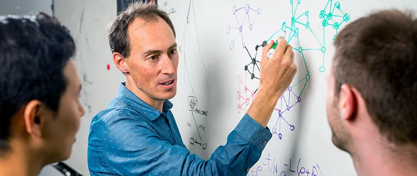Photo of Dr. Jonathan Pritchard (center) and his colleagues, Yang Li (left) and Evan Boyle, with Pritchard having drawn several interconnecting gene trees and formulas on a whiteboard in different colors.