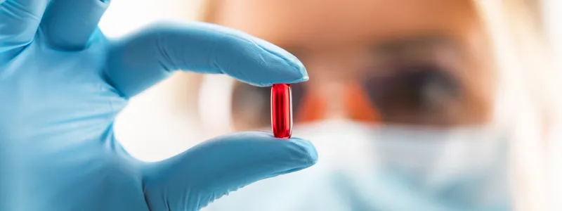 Photo of young female researcher wearing blue gloves and safety glasses, holding up a translucent red pill.