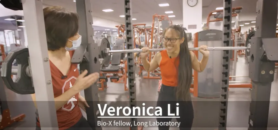 Screenshot from video of an Asian woman asking questions of an Asian female graduate stuent holding a barbell on her shoulders in a gym and smiling. A caption reads "Veronica Li, Bio-X Fellow, Long Laboratory." 
