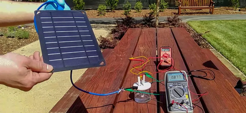 Photo of a solar panel being held up by a researcher's hands, with panel connected to a glass container and multiple sensors displaying readouts, via long wires.