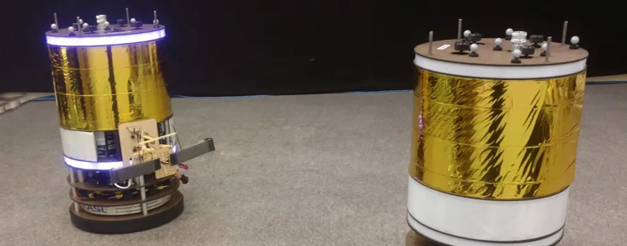 Photo of two of the space robots, which are large cylinders with gold foil around them and a ring of lights at the top.
