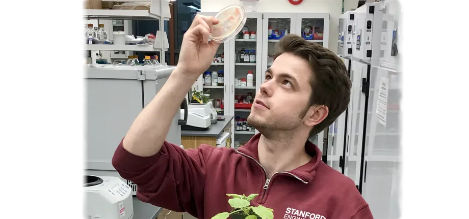 Photo of a male graduate student holding up a petri dish in one hand, with a small plant held in the other.