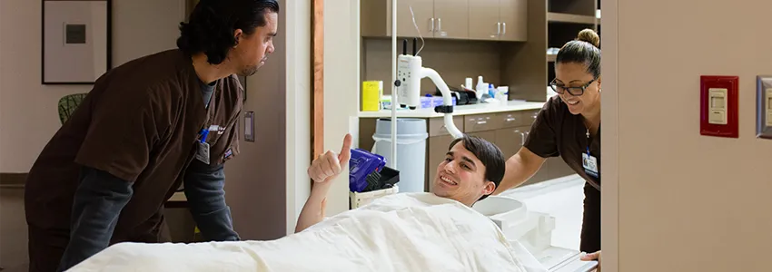 Photo of Andrew Whittaker under sheet on stretcher, giving thumbs up while being wheeled into MRI room by two nurses.